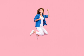 Full length photo of impressed funky girl wear blue shirt jumping high rising fists empty space isolated pink color background