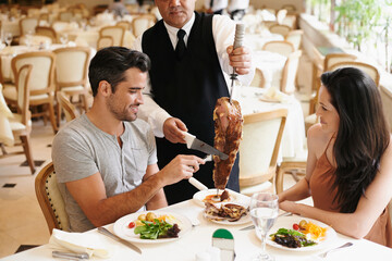 Happy couple, date and dining with kebab of waiter serving, meat or slices on romantic dinner at table. Young man or woman with chef, skewer or food for meal, eating or enjoying service at restaurant - obrazy, fototapety, plakaty