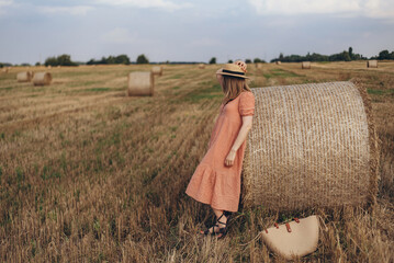 A young girl in a long dress and long hair clouded a bale of straw on a wheat field. The girl in a...