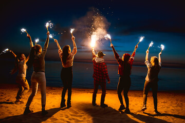Big group of happy young friends are celebrating something and having fun with candles and sparklers at sunset lake beach