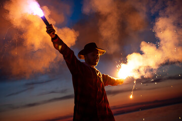 Attractive man is standing with open arms and holding bright signal fire against sunset sky