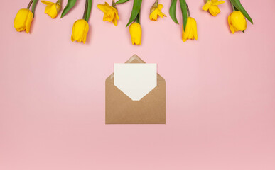 Top view of yellow tulips and daffodils on pink background. Kraft envelope, white card. Spring colourful composition, flat lay, copy space.