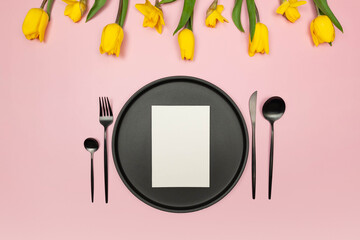 Top view of yellow tulips and daffodils on pink background. Table setting, black plate and cutlery, white card. Spring colourful composition, flat lay, copy space.
