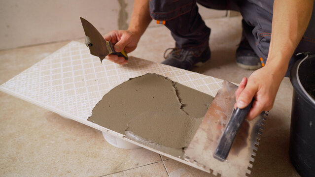 Specialist in laying ceramic tiles. Applying tile adhesive to tiles with a notched trowel - laying ceramic tiles in the bathroom. Applying tile adhesive to tiles.