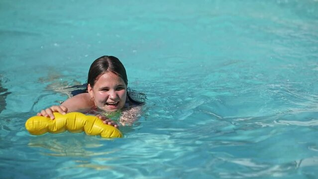 Happy teenager girl with yellow inflatable pillow in swimming pool with blue water on warm summer day on tropical vacations. Summertime activities concept.