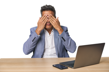 Elegant businessman at desk with laptop afraid covering eyes with hands.
