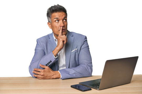 Elegant businessman at desk with laptop looking sideways with doubtful and skeptical expression.
