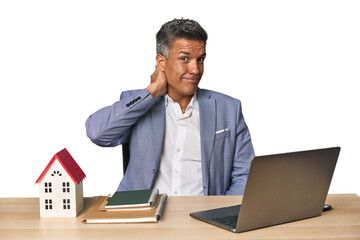 Middle-aged Hispanic businessman with real estate theme touching back of head, thinking and making...