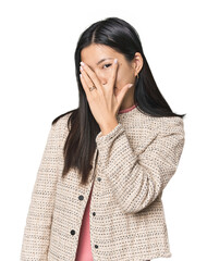 Young Chinese woman in studio setting blink at the camera through fingers, embarrassed covering face.