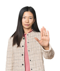 Young Chinese woman in studio setting standing with outstretched hand showing stop sign, preventing...