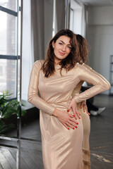 Fototapeta premium Beautiful brunette woman in long golden dress stand near mirror in room. Portrait of pretty elegant tall woman with long black hair, smile.