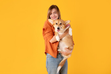 Joyful young european lady cradles her beloved cute corgi dog, both looking at camera posing against vibrant yellow background