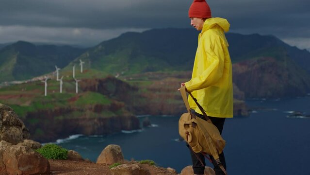 Attractive traveling woman in a yellow jacket and backpack looking at windmills in field 