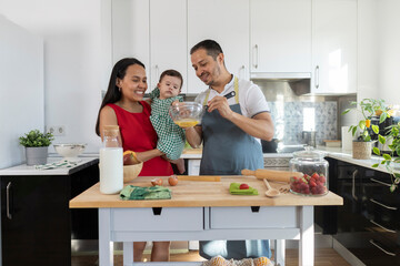 happy dad and mom cooking at home with a baby in their arms latin american family preparing food in...