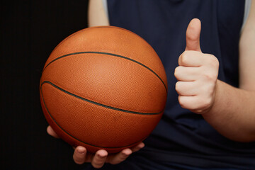basketball player’s hand holding a basketball with one hand, and the other hand showing a thumbs up. Sports victory in basketball, invitation to a basketball match