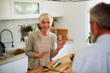 A smiling senior woman talking with her husband in the kitchen while slicing a cucumber for a salad