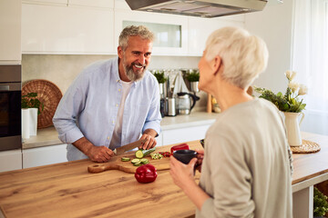 A happy senior adult man talking to his wife in the kitchen and having a laugh while preparing a...
