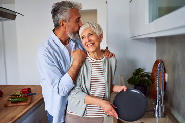 A loving senior adult husband embracing his wife and planting a kiss to the top of her head while she is washing the dishes