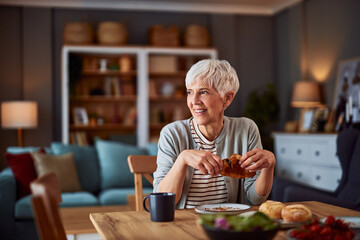 A smiling senior adult woman day dreaming and enjoying a delicious croissant for breakfast at the...