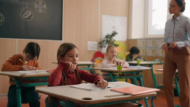 Caucasian woman teacher female tutor walking between classroom tables desk in elementary school teach children help with test task check exercise in notebooks class kids writing teach studying lesson