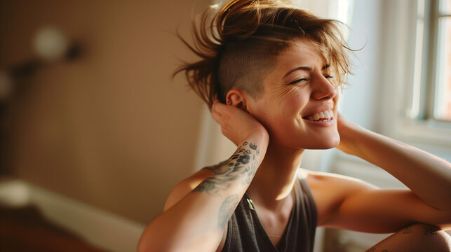 Smiling queer woman sitting, touching hair, tattoo visible, sunlit room.