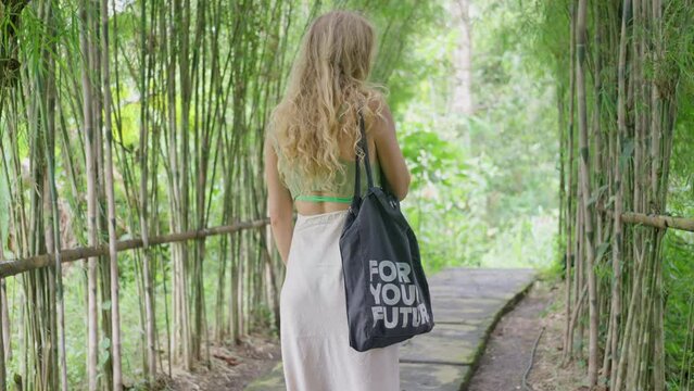 A woman is walking down a path in the woods with a drawstring bag. A Girl with long hair is strolling through the woods, carrying a drawstring bag slung over her waist. Slow motion