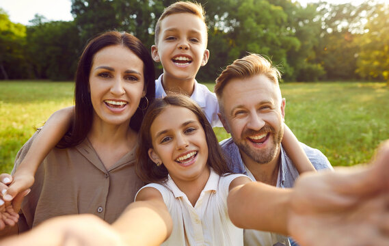 Selfie portrait of a happy smiling family of four in nature. Parents with kids spending time together and having fun in summer park. Little girl taking photo with mother, father and brother.