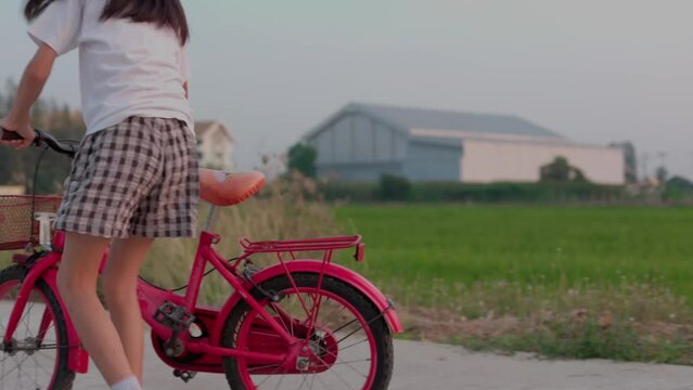 Video taken using a handheld camera Portrait Asian Thai kid girl, aged 8 to 10 years old, riding a red bicycle, playing on the road outdoors, happy and having fun. The background is a green field.