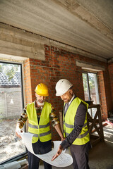 cheerful cottage builder and architect and with safety gear working with blueprint on site - 742713037