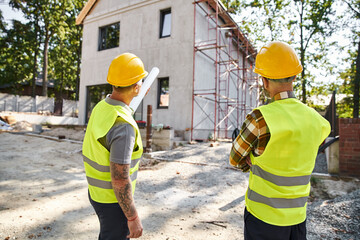 back view of devoted cottage builders in safety helmets with blueprint discussing their work on site