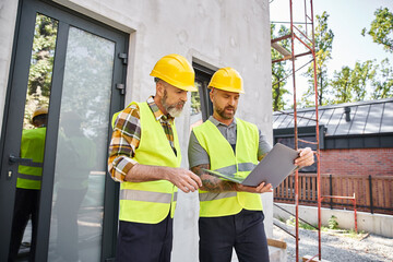 handsome bearded men in safety vests and helmets working with laptop on construction site, builders