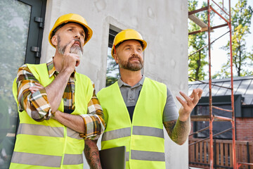two good looking construction workers in safety vests and helmets discussing site, cottage builders