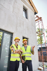 two good looking construction workers in safety vests and helmets discussing site, cottage builders