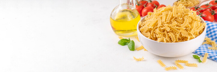 Raw uncooked Mafaldine pasta with ingredients for cooking. Short italian Mafaldine pasta with olive oil, cherry tomatoes, basil, spices on white kitchen table, background copy space