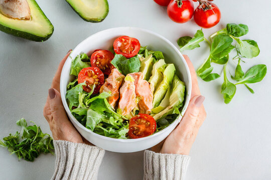 Woman's hands holds a white bowl with salad and tomatoes,avocado, chicken, green leaves,healthy eating concept.white background.Top view.