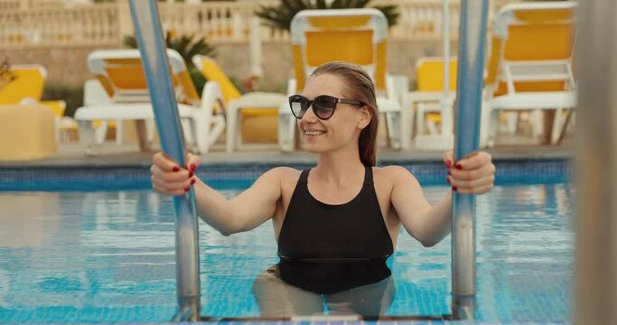 attractive smiling woman wearing black one piece swimsuit and sunglasses standing on resort hotel swimming pool stairs