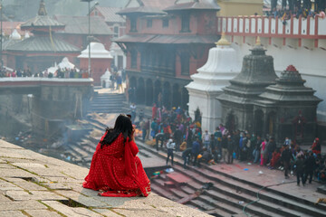 Back view of woman in red sari watching mournful cremation ceremony at Pashupatinath Temple...