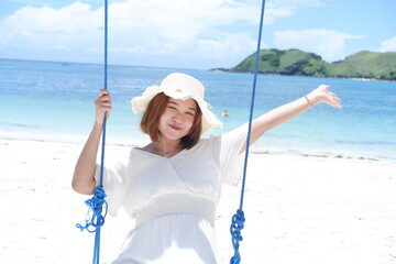 Woman in white dress and hat swinging on tropical beach, sunny day, good weather on lombok beach