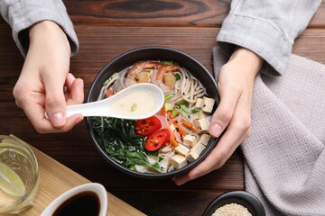 Woman eating delicious ramen with spoon at wooden table, top view. Noodle soup