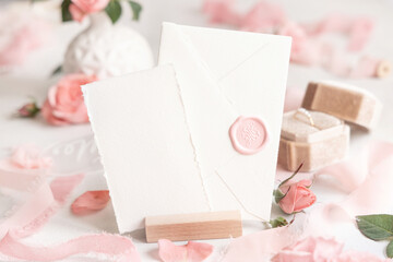Card and sealed envelope near pink roses, petals and silk ribbons close up, wedding mockup