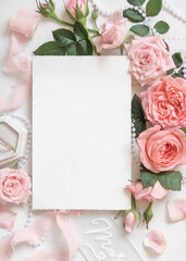 Vertical blank card near pink roses, engagement ring and silk ribbons top view, wedding mockup