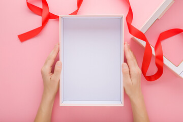 Young woman hands holding opened empty white present box with red ribbon on pink table background....