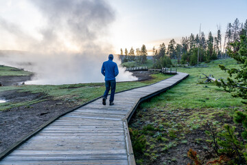 A man, visitor walking down an elevated wooden boardwalk over grassy land with hot springs steaming...