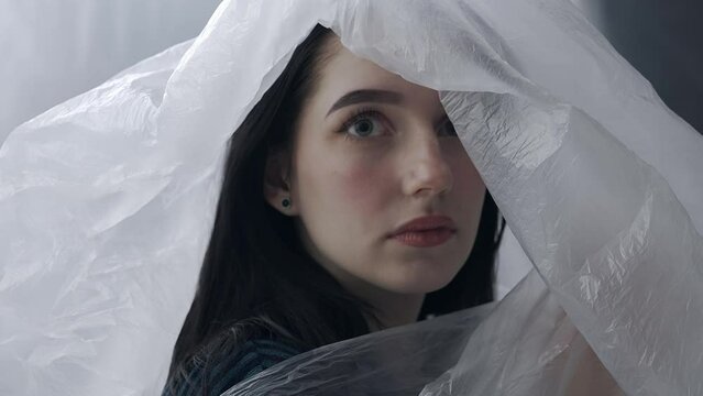 Female in sleepwear posing in the studio. Young woman in pajama shirt posing at camera under thin sheet of oilcloth wrap.