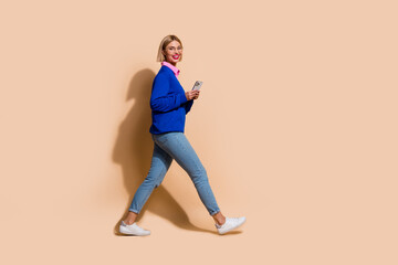 Fototapeta na wymiar Full length profile portrait of pretty cheerful person hold use smart phone walk empty space isolated on beige color background