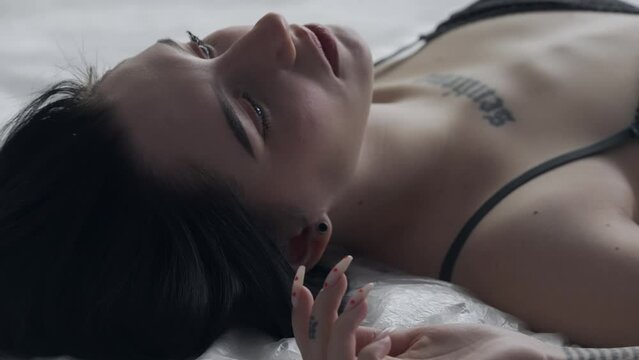 Female in underwear laying on the bed in the studio. Young woman seminude in lingerie laying posing under thin sheet of oilcloth wrap, close up.