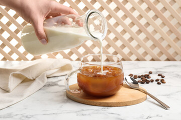 Woman pouring milk into glass with refreshing iced coffee at white marble table, closeup
