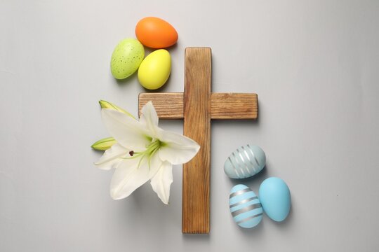 Wooden cross, painted Easter eggs and lily flowers on grey background, flat lay