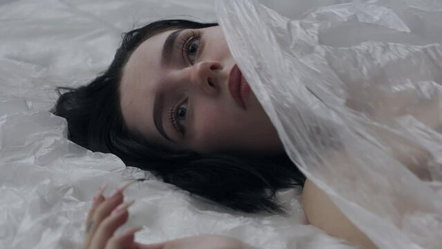 Female in underwear laying on the bed in the studio. Young woman seminude in lingerie laying posing under thin sheet of oilcloth wrap, staring at camera.