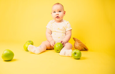 Cute baby girl with green apples sitting in studio on yellow background.